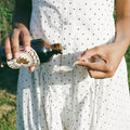 Person in a white polka dot dress pouring a dark liquid from a bottle into a small container outdoors.