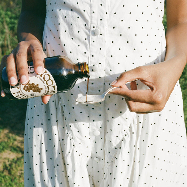 Person in a white polka dot dress pouring a dark liquid from a bottle into a small container outdoors.